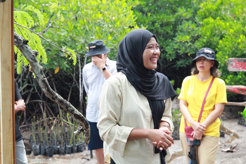 Seorang perempuan berhijab hitam mengenakan kemeja krem tersenyum saat kegiatan luar ruang, dengan peserta lain berdiri di latar belakang di area hijau pesisir.