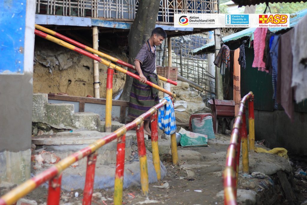A man walks down a pathway in a camp area, using red and yellow bamboo handrails for support. Clothes hang on a line nearby, and makeshift houses with corrugated walls surround the path.