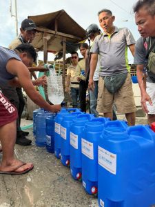 Several men and women are gathered around a line of blue plastic water containers labelled “ECT WASH.” One man is crouching and filling a large container with water from a hose connected to a water trucking supply. Others stand nearby, waiting and observing. The setting is an outdoor roadside area with simple wooden structures in the background. The activity supports households affected by the earthquake in Barangay San Vicente, Bogo, where the community relies on trucked water due to power outages affecting their regular water supply.