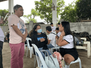 Several people are gathered in an outdoor area for a rapid assessment activity. A woman wearing a face mask and an A2D shirt sits on a white plastic chair, listening attentively. Next to her, a woman with a lower limb amputation sits with a pink bag on her lap, actively engaging in the discussion. A man stands nearby, observing the conversation. Other participants are seated in the background. The setting suggests an interview process to identify the needs of people affected by the earthquake, including persons with disabilities.