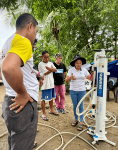 A woman wearing a black hat with ACCORD logo and a white polo shirt stands next to a Skyhydrant unit, demonstrating how to set it up. She gestures with her hands while several A2D members and community members stand around her, attentively observing the process. The setup includes hoses connected to the Skyhydrant, and the activity takes place outdoors under trees, indicating a hands-on technical demonstration session.