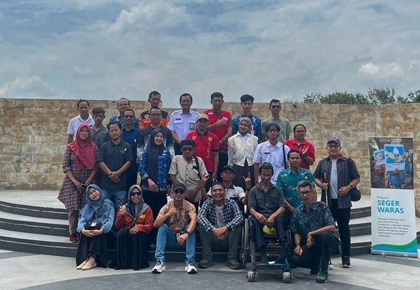 Foto bersama 28 partisipan dan penyelenggara Program Seger Waras, terdiri dari laki-laki dan perempuan dengan latar belakang beragam, termasuk perwakilan pemerintah daerah, organisasi masyarakat sipil, desa dampingan, serta individu dengan disabilitas. Mereka berpose di area terbuka dengan latar dinding batu alam dan langit cerah. Barisan depan duduk di tangga, termasuk peserta pengguna kursi roda, sementara barisan tengah dan belakang berdiri rapat. Beberapa peserta mengenakan seragam putih, kemeja batik, pakaian kasual, hingga seragam organisasi. Di sisi kanan foto terdapat standing banner bertuliskan “Program Seger Waras” yang menandakan kegiatan terkait peningkatan kapasitas lokal, kesehatan lingkungan, dan pengelolaan risiko bencana. Ekspresi peserta sebagian besar tersenyum.