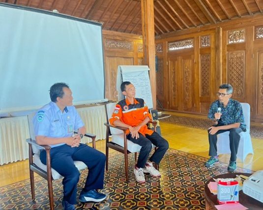 Peserta dan fasilitator duduk membentuk setengah lingkaran di tengah ruangan. Fasilitator sambil memegang mikrofon untuk bertanya kepada peserta. Di sisi kanan tampak perangkat seperti LCD yang digunakan untuk menampilkan data dari yang disampaikan oleh peserta. Peserta dalam podcast ini terdiri dari 2 perwakilan. Peserta menggunakan baju warna putih merupakan perwakilan dari BMKG (Badan Meteorologi, Klimatologi, dan Geofisika), sedangkan peserta yang menggunakan baju warna orange merupakan Sekretaris Desa Giritengah.