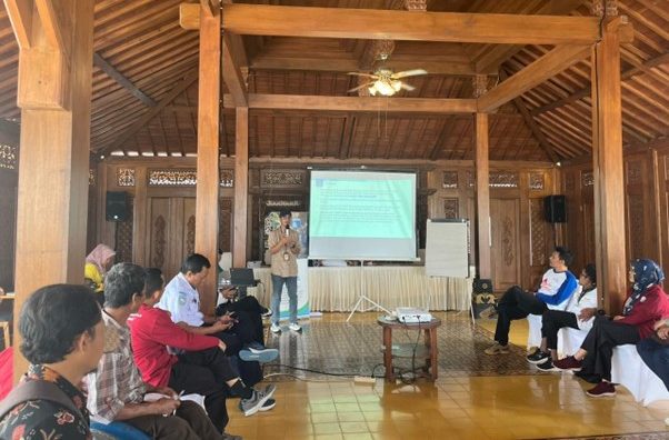 Fasilitator berdiri di depan sambil menampilkan materi di layar. Sementara peserta duduk membentuk letter U untuk mendengarkan fasilitator. Suasana penyampaian materi tersebut bertujuan agar peserta bisa tetap fokus dan interaktif.