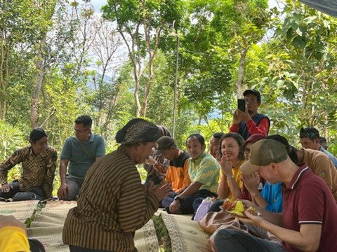 Alt Teks: dengan senyum ramah, seorang laki-laki mengenakan busana lurik khas Jawa memberikan potongan pertama tumpeng kepada Martin dari Jerman sebagai bentuk simbolis.