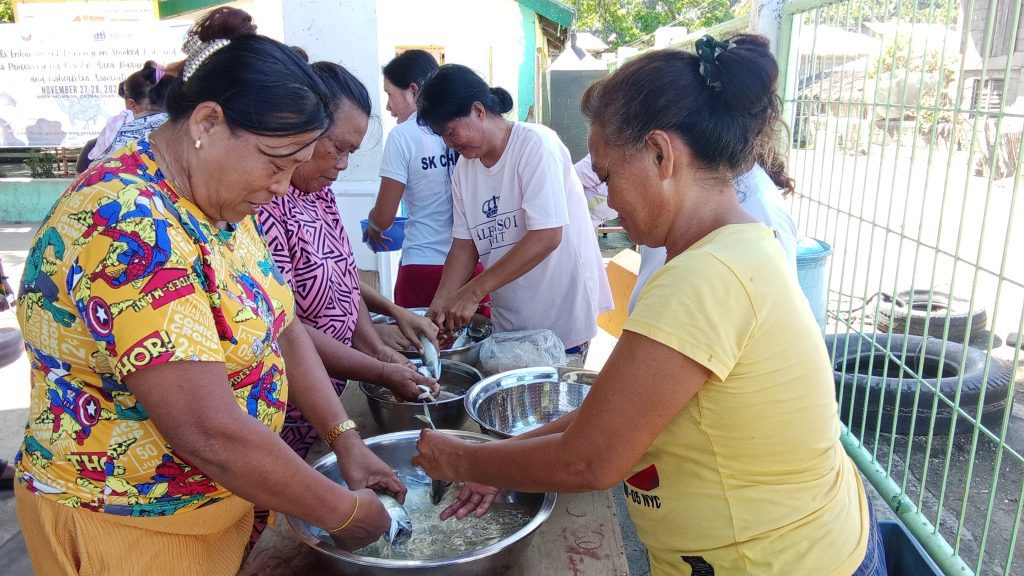 Several women from the Mombon Women’s Organisation work together outdoors, cleaning and preparing fish in large metal bowls during a practical fish processing training session.