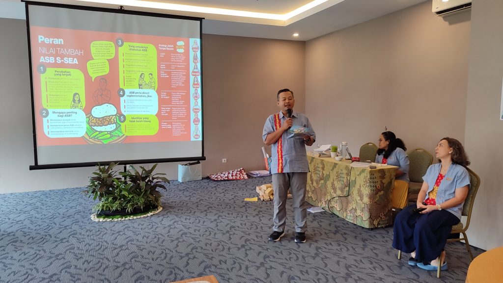 An ASB S-SEA team member speaking during a presentation, with colleagues seated nearby and a presentation on ASB’s added value projected on the screen.