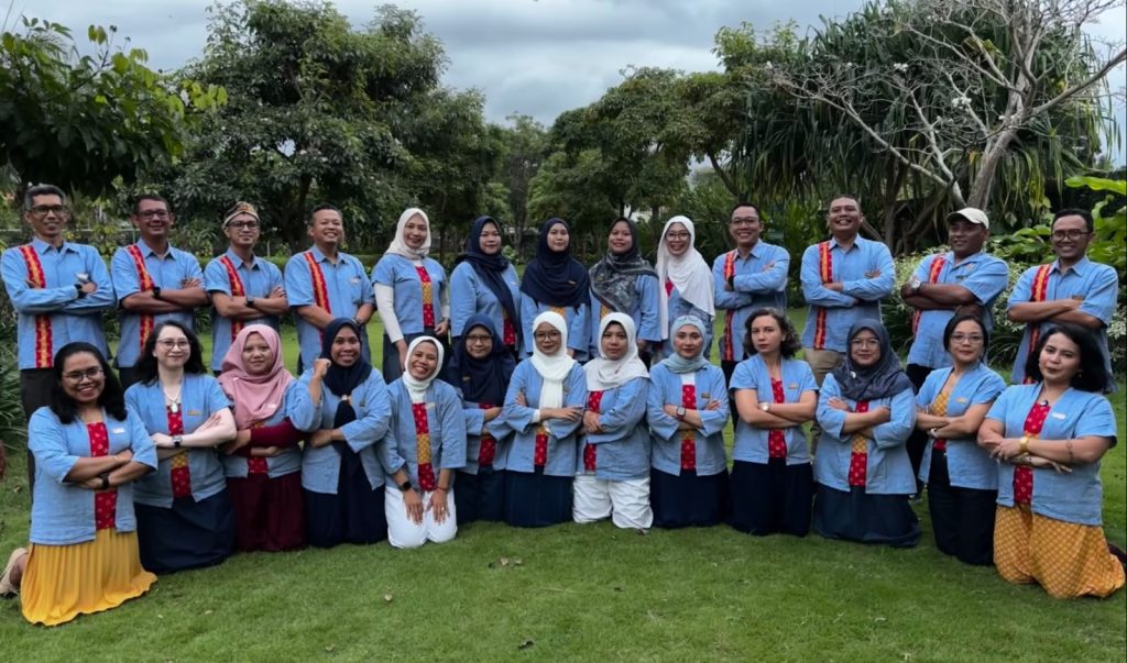 Group photo of ASB S-SEA team members standing and kneeling together on a lawn, wearing matching light blue shirts with red patterned accents, specifically batik which is local indonesia accents.
