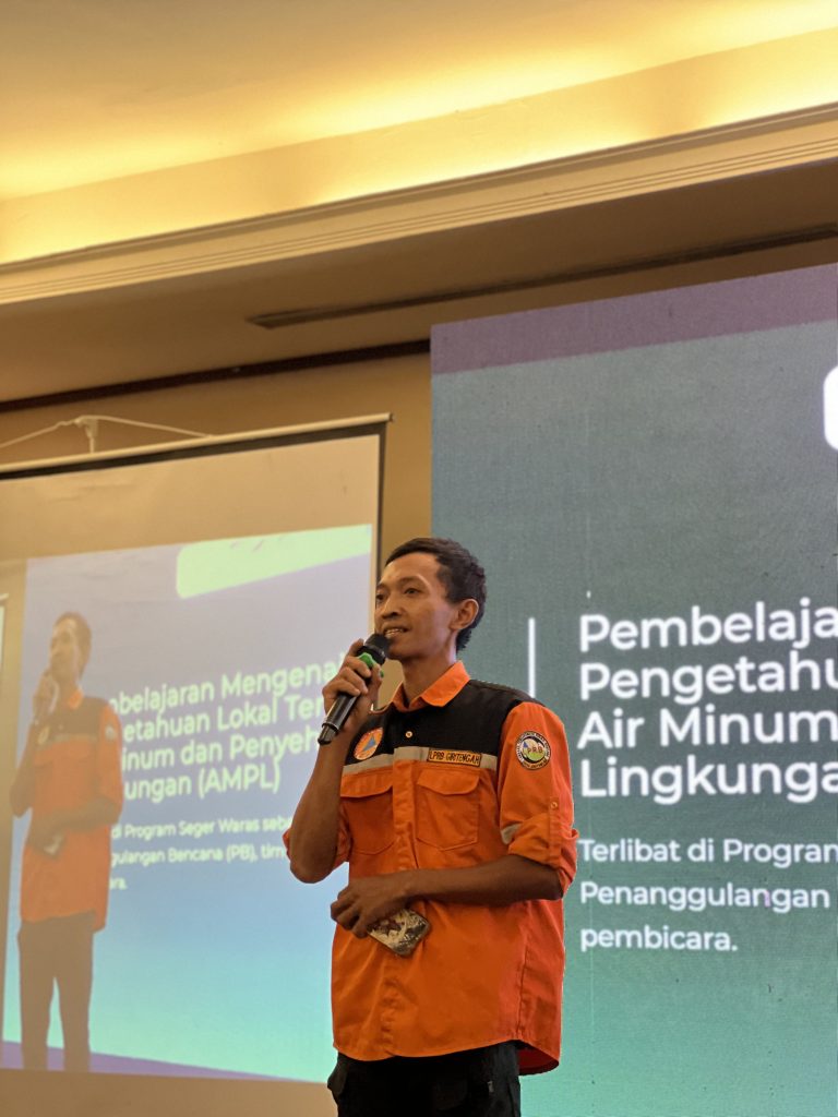 Mas Lekan berbicara menggunakan mikrofon saat menyampaikan presentasi, mengenakan seragam oranye penanggulangan bencana, dengan slide presentasi terpampang di layar di belakangnya.