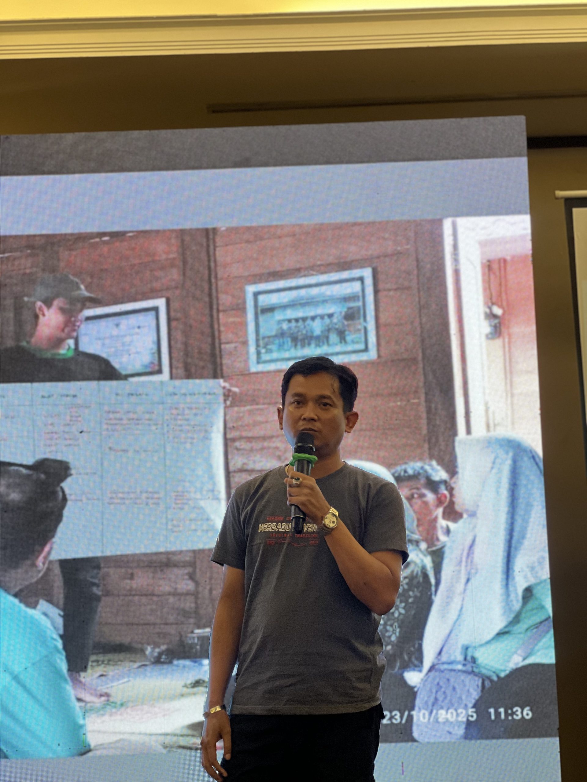 Pak Alfonso berbicara menggunakan mikrofon saat menyampaikan presentasi, mengenakan kaos berwarna abu-abu, dengan slide presentasi terpampang di layar di belakangnya.