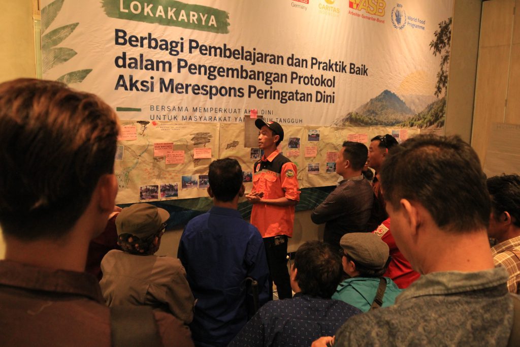 A group of people gathering in front of a workshop banner while a man in an orange uniform and cap presents, pointing toward a board covered in notes and photos.