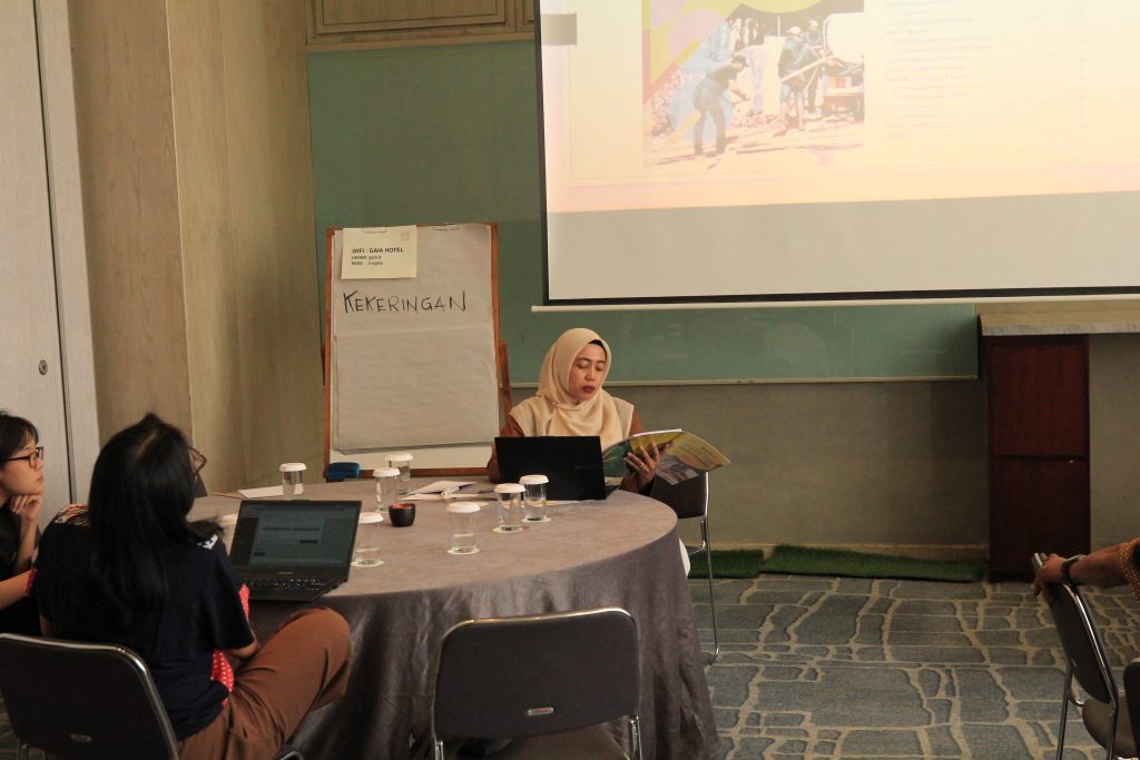 A woman in a hijab sits at a round table, reading a document next to her laptop during a workshop, while other participants listen. A flip chart with the word "KEKERINGAN" (Drought) and a large projector screen are visible in the background.