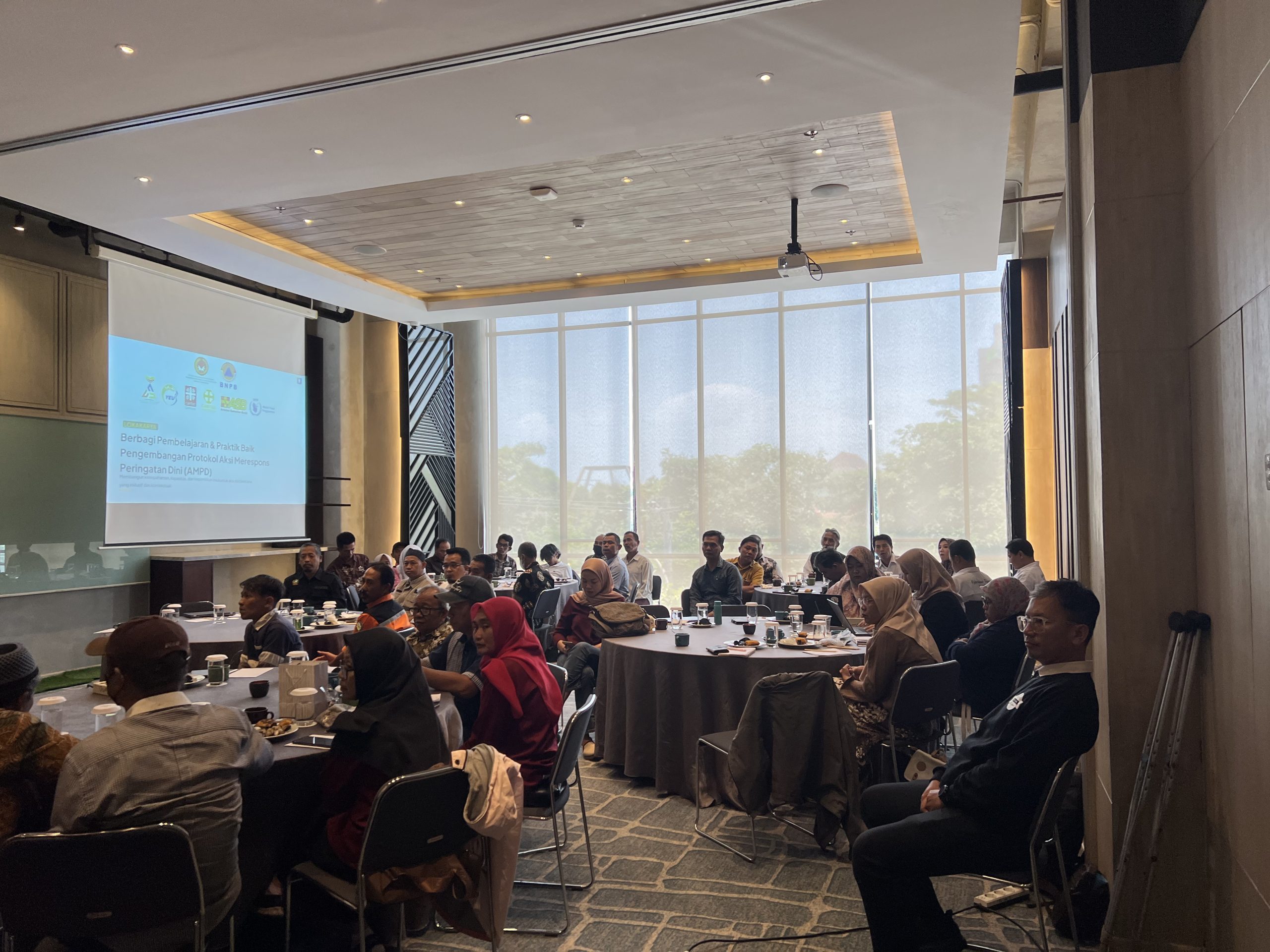 A meeting room where participants are seated at round tables facing a projector screen displaying a presentation on the "Workshop on Sharing Lessons Learnt and Good Practices in Developing Anticipatory Action Protocols".