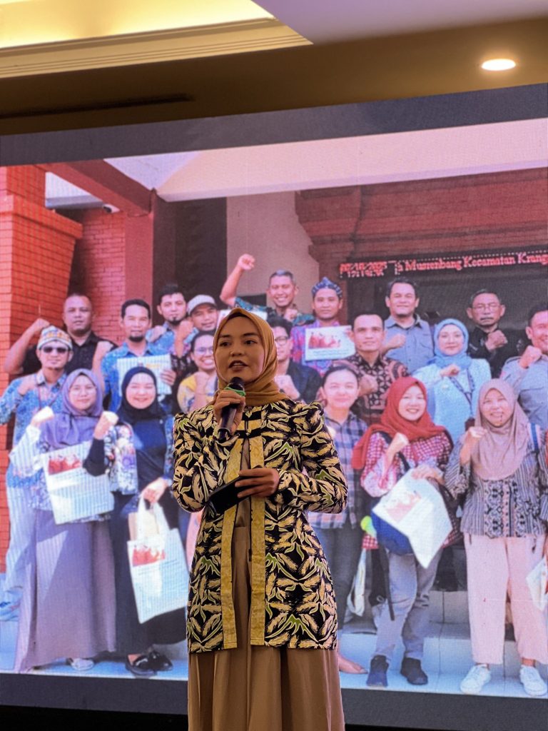 Perempuan dengan nuansa batik bernama bu Wahyu sedang berbicara dalam sesi berformat TED Talk dengan memegang microphone dan presentasi di belakangnya.